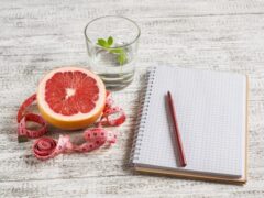 Open food diary next to a grapefruit, glass of water, and measuring tape