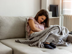 Unhappy woman holding hands on stomach suffering from abdominal pain with eyes closed. Women having menstrual period, food poisoning, gastritis or diarrhea. Girl feeling unwell sitting in living room.
