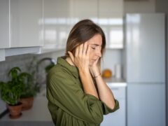 Tired woman feeling headache rubbing temples standing on kitchen with closed eyes at home. Blonde female suffering from migraine head pain. Having headache cephalalgia premenstrual syndrome feel bad.