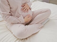 A young hispanic woman sits on a bed, cradling her pregnant belly in a cozy bedroom, evoking maternity serenity.
