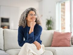 Happy relaxed 50 years old woman sitting on sofa at home looking away. Smiling middle aged lady posing in living room at home. Mature older female with curly gray hair relaxing on couch enjoying chill