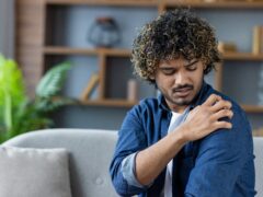 A person appears to be experiencing discomfort and scratching their shoulder while indoors. The scene is cozy and well-lit, with bookshelves and plant decor visible in the background.